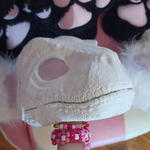 Op de afbeelding: Een wit dinosaurusmasker met een getextureerd oppervlak, met grote oogopeningen. Het masker wordt vastgehouden door een persoon die meerdere roze en witte kralenarmbanden draagt. Zwart-witte pluche poten in de vorm van poten staan op de achtergrond.