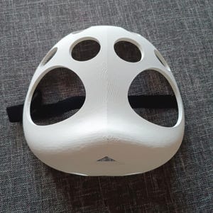 以下が含まれることがあります： 複数の丸い穴と黒いストラップが付いた白いプラスチック製のマスク。マスクは鼻と口を覆うように設計されています。