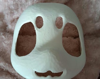 May include: A white, blank mask with large eye and mouth openings. The mask has a smooth, rounded shape and is likely intended for costume or theatrical use. The mask is resting on a pink, textured surface.