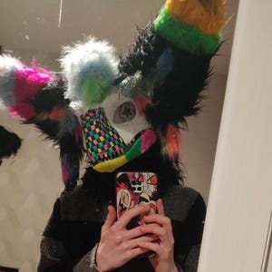 May include: A colorful furry mask with large, fluffy ears in rainbow hues, including pink, yellow, and blue. The mask has a white face with green and black accents. The person is holding a phone with a cartoon design.