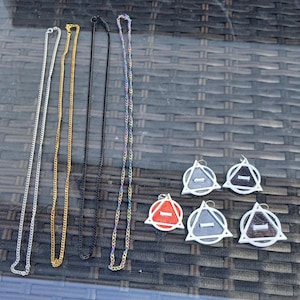 Peut inclure: Un assortiment de colliers et de boucles d'oreilles. Les colliers sont argentés, dorés, noirs et irisés. Les boucles d'oreilles sont blanches avec un motif triangulaire et un centre coloré, dont le rouge, le gris et le noir.