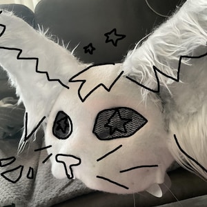 Op de afbeelding: Een wit imitatiebont dierenkopmasker met grote, puntige oren en mesh oogbedekkingen. Het masker is versierd met zwarte handgetekende contouren, waaronder stervormen in de ogen, snorharen en grillige lijnen langs de randen van de oren.