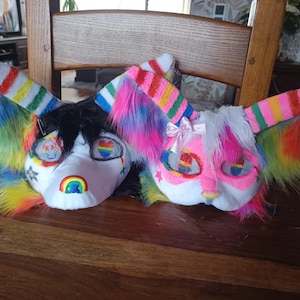 May include: Two colourful furry animal masks with rainbow ears and accents. One mask is white with black fur and the other is pink with white fur. Both masks have rainbow-coloured accents and are decorated with hearts and rainbows.