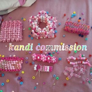 Könnte beinhalten: Eine Auswahl handgefertigter Kandi-Schmuckstücke in Rosa-, Weiß- und anderen Farbtönen. Die Stücke umfassen Armbänder, Manschetten und ein sternförmiges Design, alle aus bunten Perlen. Der Text "kandi commission" ist ebenfalls sichtbar.