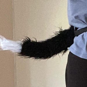 Op de afbeelding: Een accessoire in de vorm van een dierenstaart van imitatiebont met een lange, zwarte, ruige textuur en een opvallende witte punt. De staart is bevestigd aan een zwarte stoffen riem die om de taille wordt gedragen en horizontaal naar achteren uitsteekt.