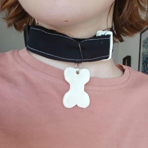 Könnte beinhalten: Ein schwarzes Choker-Halsband mit einem weißen knochenförmigen Anhänger. Das Halsband besteht aus schwarzem Stoff mit weißen Nähten und einem weißen Verschluss. Der Knochenanhänger ist weiß und hängt an einer dünnen schwarzen Schnur.