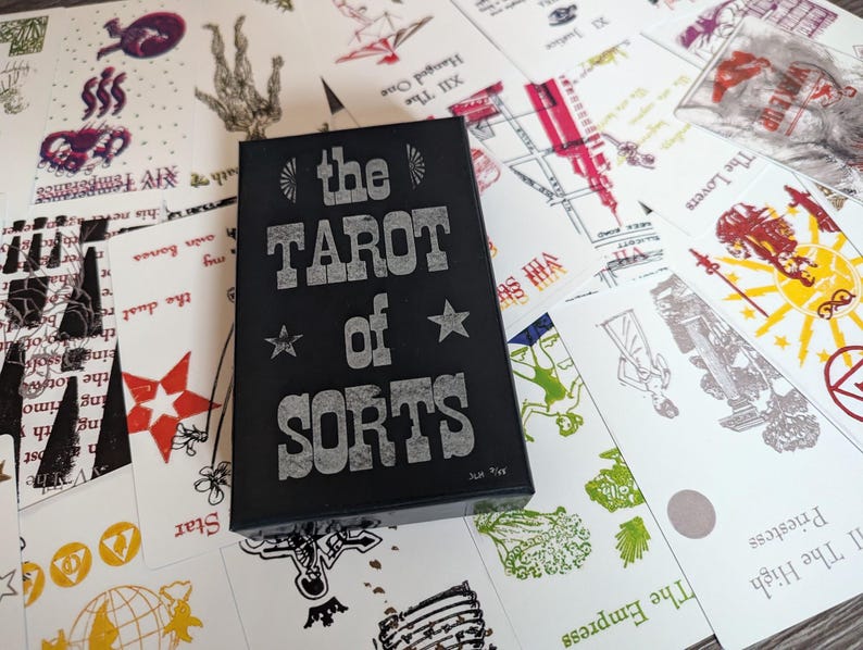 Może przedstawiać: Czarne pudełko z napisem "The Tarot of Sorts" srebrnymi literami. Pudełko jest otoczone kartami tarota z r&oacute;żnymi obrazami i tekstami.
