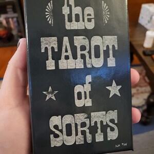 Może przedstawiać: Czarne pudełko z białym tekstem, kt&oacute;ry brzmi "the TAROT of SORTS" z dwiema białymi gwiazdami i stylizowanym wzorem słońca. Tekst "JLH 7/63" znajduje się w prawym dolnym rogu.