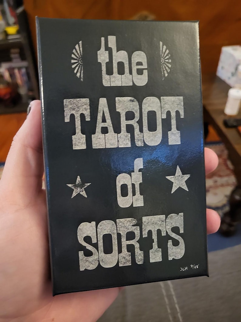 Może przedstawiać: Czarne pudełko z białym tekstem, kt&oacute;ry brzmi "the TAROT of SORTS" z dwiema białymi gwiazdami i stylizowanym wzorem słońca. Tekst "JLH 7/63" znajduje się w prawym dolnym rogu.