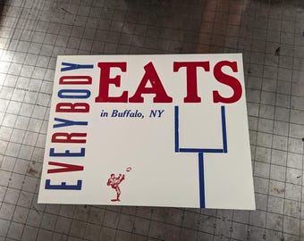 Może przedstawiać: Biały prostokątny znak z napisem "EVERYBODY EATS" w czerwonych i niebieskich literach blokowych. Tekst "in Buffalo, NY" znajduje się pod słowem "EATS". Piłkarz kopie piłkę w lewym dolnym rogu.