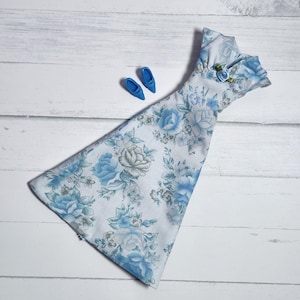May include: A sleeveless doll dress featuring a white fabric base with a repeating blue floral pattern, a high neckline, and a small blue fabric rose accent at the collar, accompanied by a pair of matching blue plastic doll shoes.