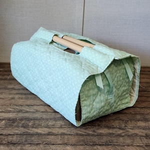 May include: A rectangular quilted fabric carrier in light mint green with a small white floral pattern. The carrier features two light-colored wooden dowel handles at the top and matching mint green ribbon accents on the side panels.