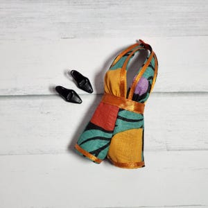 May include: A sleeveless doll dress featuring a vibrant abstract pattern in teal, mustard yellow, red, and purple, accented with orange satin trim at the neckline, armholes, and waist, paired with a set of small black pointed-toe doll shoes.