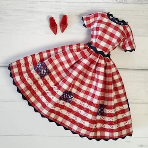 May include: A short-sleeved dress featuring a red and white gingham pattern with a dark blue scalloped trim at the neckline, waist, and hem. The skirt includes decorative white cross-stitching and blue lattice embroidery. Two red pointed-toe shoes.