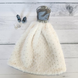 May include: A doll-sized formal dress with a silver-trimmed bodice and a fluffy, cream-colored skirt sprinkled with silver sequins. Matching silver doll shoes and earrings are also present, creating a miniature ensemble.