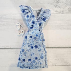 May include: A light blue floral print dress with a V-neck and ruffled sleeves. The dress is paired with white high-heeled sandals. The dress features a wrap-style design and a matching belt. The floral pattern includes dark blue flowers and leaves.