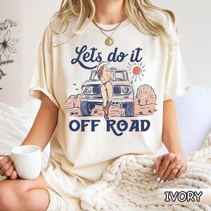 May include: Ivory-colored t-shirt with a graphic of a woman in a cowboy hat leaning against a vintage off-road vehicle. The text "Lets do it OFF ROAD" is printed above and below the image. The shirt is a relaxed fit.