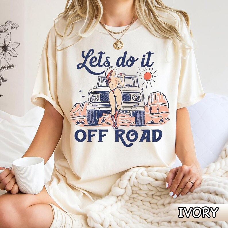 May include: Ivory-colored t-shirt with a graphic of a woman in a cowboy hat leaning against a vintage off-road vehicle. The text "Lets do it OFF ROAD" is printed above and below the image. The shirt is a relaxed fit.