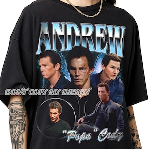 May include: Black t-shirt with a graphic design featuring the name "ANDREW" in a blue and white font. The design includes multiple images of a person, with the text "DONT COPY MY DESIGN" and "Pope Cody".