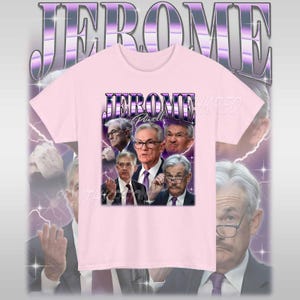 May include: Pink t-shirt with a graphic print of Jerome Powell, the chairman of the Federal Reserve, in a retro style. The graphic features Powell's face surrounded by lightning bolts and the text "JEROME Powell" in a bold, purple font.