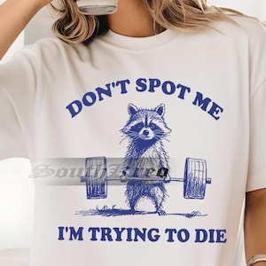 May include: White t-shirt with a blue graphic of a raccoon lifting a barbell. The text "Don't Spot Me I'm Trying To Die" is printed on the shirt.