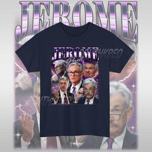 May include: A navy blue t-shirt with a graphic print of Jerome Powell, the current chairman of the Federal Reserve, in a retro style. The text "Jerome Powell" is printed in purple on the shirt.