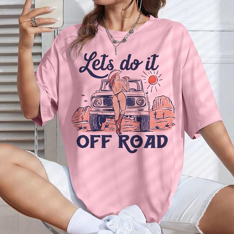 May include: Pink t-shirt with the text "Lets do it OFF ROAD" and a graphic of a woman in a cowboy hat leaning against a vintage off-road vehicle. The design includes a desert landscape with a sun.