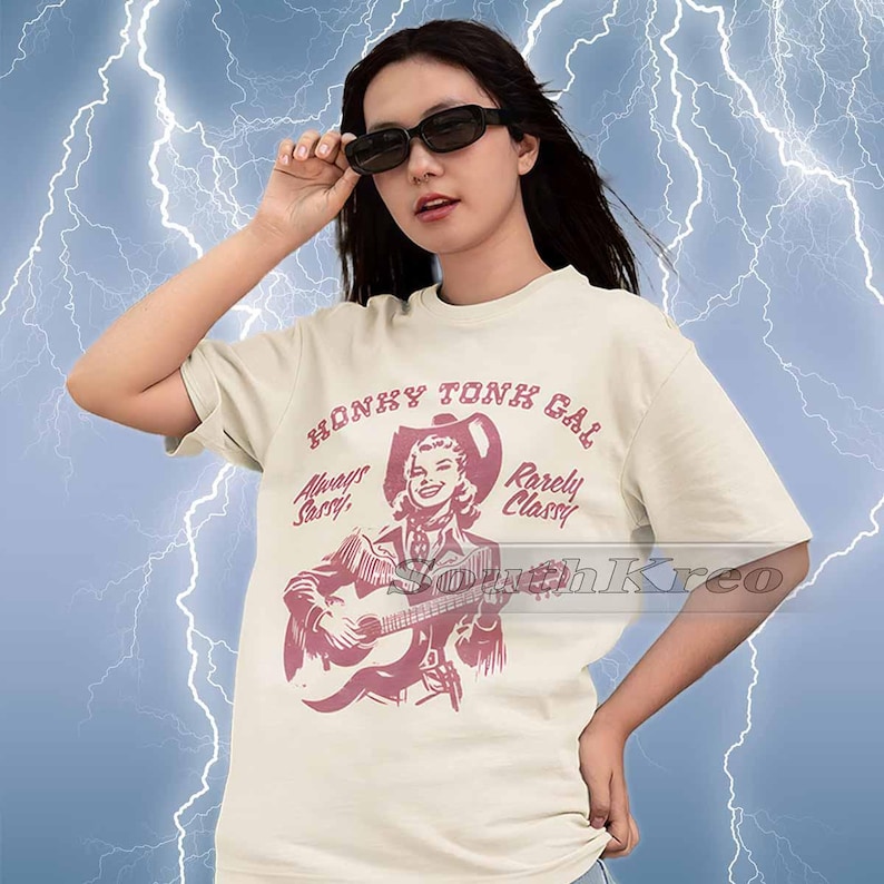 May include: A white t-shirt with a vintage-style illustration of a woman playing a guitar. The text on the shirt reads "Honky Tonk Gal, Always Sassy, Rarely Classy".