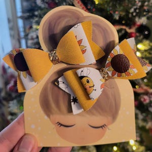 May include: Two yellow hair bows with sunflower and bee designs. The bows have a glittery center and are attached to a card with a cartoon illustration of a girl. The bows are yellow, white, and brown.