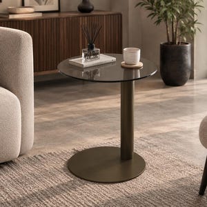 Può includere: Un tavolino rotondo con piano in vetro fumé scuro, sostenuto da una colonna cilindrica centrale e una base circolare piatta, entrambi rifiniti in color bronzo scuro opaco. Sul piano del tavolo sono appoggiati una tazza in ceramica e un libro.