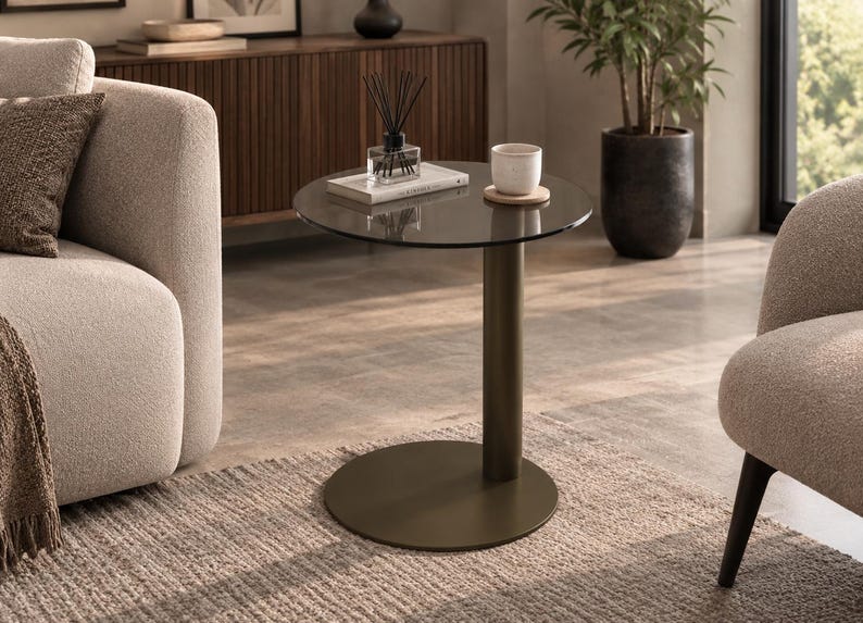 Op de afbeelding: Een ronde bijzettafel met een donker getint glazen blad, ondersteund door een enkele cilindrische kolom en een platte ronde voet, beide afgewerkt in een matte donkerbronzen kleur. Op het tafelblad staan een witte keramische beker en een boek.
