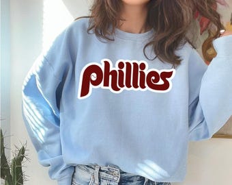 May include: A light blue sweatshirt with the word "Phillies" in maroon script across the chest. The sweatshirt is a crew neck style and appears to be made of a soft material. The text has a white outline.