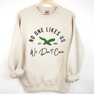 Könnte beinhalten: Ein beiger Pullover mit dem Text "No One Likes Us Est. 1933 We Don't Care" in Schwarz. Ein grüner Adler befindet sich über dem Text.