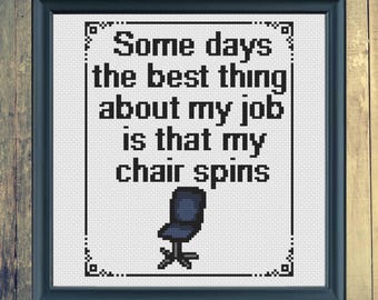 Puede incluir: Obra de arte en punto de cruz enmarcada con el texto "Some days the best thing about my job is that my chair spins". El diseño incluye una imagen pixelada de una silla de oficina. El marco es azul oscuro, sobre un fondo de madera.