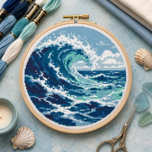 May include: A circular wooden embroidery hoop containing a cross-stitch pattern of a large ocean wave. The design features shades of navy, royal blue, teal, and white, depicting a crashing wave with sea foam against a light blue sky background.