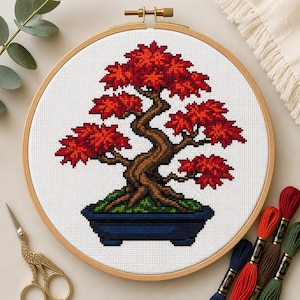 May include: A cross-stitch embroidery project in a round wooden hoop featuring a bonsai tree with a brown trunk and vibrant red leaves, planted in a dark blue rectangular pot, set against a white fabric background with a grid pattern.