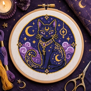 Puede incluir: Un bastidor de bordado circular de madera que contiene un patrón de punto de cruz de un gato azul oscuro con marcas celestiales doradas, incluyendo una luna creciente en la frente y el cuerpo, rodeado de nubes moradas y estrellas.