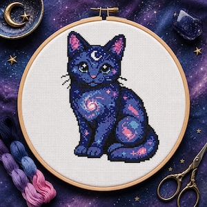 Könnte beinhalten: Ein Kreuzstich-Stickprojekt in einem runden Holzrahmen, das eine dunkelblaue Katze mit Galaxienmuster, rosa Innenohren und einer weißen Mondsichel auf der Stirn zeigt, auf einem weißen Stoffhintergrund mit sichtbaren Kreuzstich-Gittern.