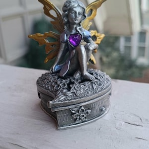 May include: A silver-toned fairy figurine with gold wings and a purple heart-shaped gem. The fairy sits atop a heart-shaped box with floral detailing. The wings are gold and the fairy is wearing a blue dress.