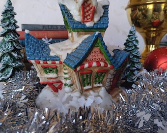 Puede incluir: Una figura de cerámica de Santa's Ski Lodge con techo azul, adornos rojos y blancos, y el texto "SANTA'S SKI LODGE". El albergue está rodeado de oropel plateado y pequeños árboles de Navidad verdes. Una bola roja y un objeto dorado están en el fondo.