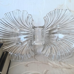 May include: Clear glass serving dish with a butterfly or bow tie shape. The dish has a ribbed design on the wings and three small circular details on each side. The dish is on a light background.