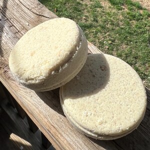 May include: Two round, light beige bath bombs with a textured surface are displayed on a weathered wooden surface. The bath bombs are circular and appear to be stacked, with a visible seam. The background shows green grass and sunlight.