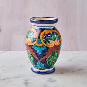 May include: A hand-painted ceramic vase with a dark blue base and colorful floral designs. The vase features orange, green, and blue accents, with a white and blue striped rim. The vase is approximately 7 inches tall.