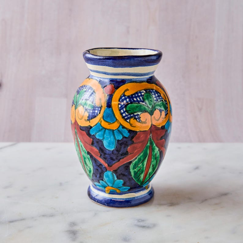 May include: A hand-painted ceramic vase with a dark blue base and colorful floral designs. The vase features orange, green, and blue accents, with a white and blue striped rim. The vase is approximately 7 inches tall.