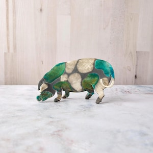 May include: A decorative pig figurine featuring a mottled pattern of dark gray, cream, and vibrant teal green patches. The animal is depicted in a standing position with its head lowered, showcasing a smooth, glossy finish across its entire body.