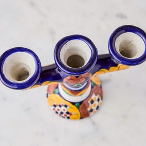 May include: A handcrafted, ceramic candle holder with three openings. The candle holder features a vibrant design with blue, yellow, and red accents. The base is round, and the openings are surrounded by a dark blue rim.