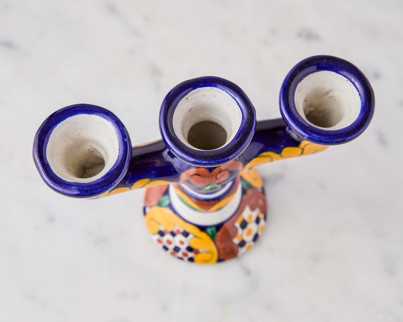 May include: A handcrafted, ceramic candle holder with three openings. The candle holder features a vibrant design with blue, yellow, and red accents. The base is round, and the openings are surrounded by a dark blue rim.