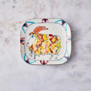 May include: Square ceramic plate with a hand-painted sheep design. The sheep has a white face, orange horns, and a colorful body with yellow, red, and orange accents. The plate's border features a geometric pattern with blue, red, and purple triangles.