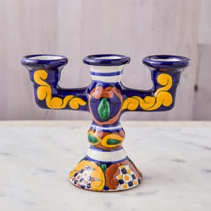 May include: A hand-painted ceramic candle holder with three candle cups. The candle holder is primarily dark blue with yellow, orange, and green floral designs. The base is flared, and the overall design is symmetrical.