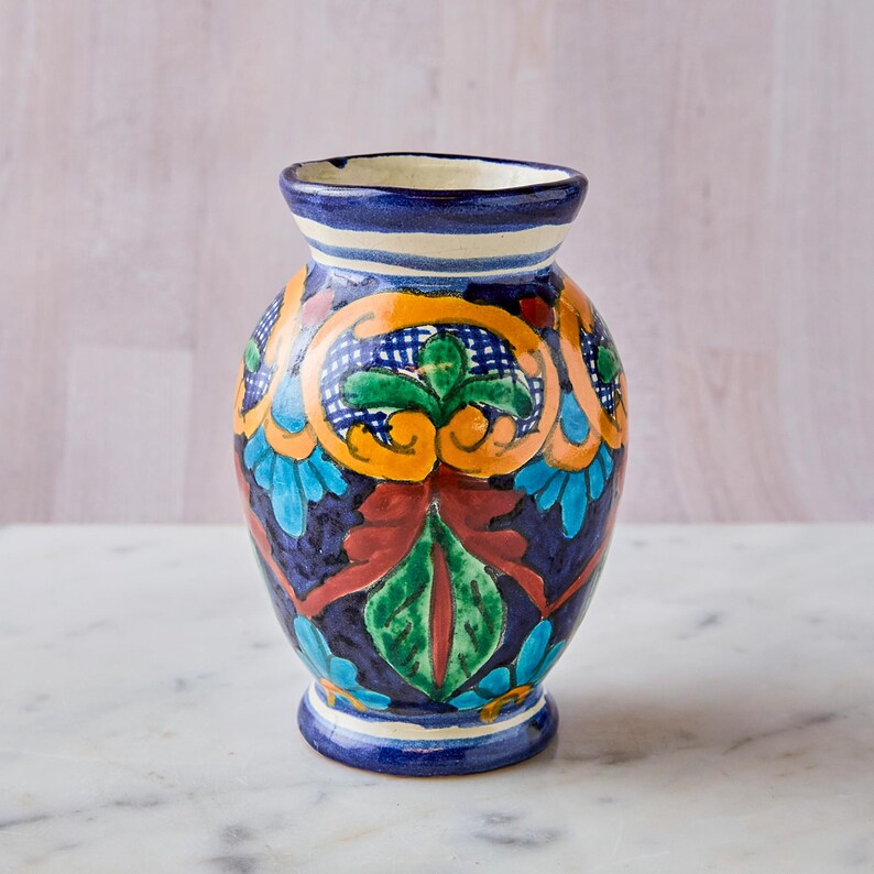 May include: A hand-painted ceramic vase with a vibrant design. The vase features a dark blue base with colorful floral and geometric patterns in orange, green, red, and blue. The rim is dark blue with a white and light blue stripe.